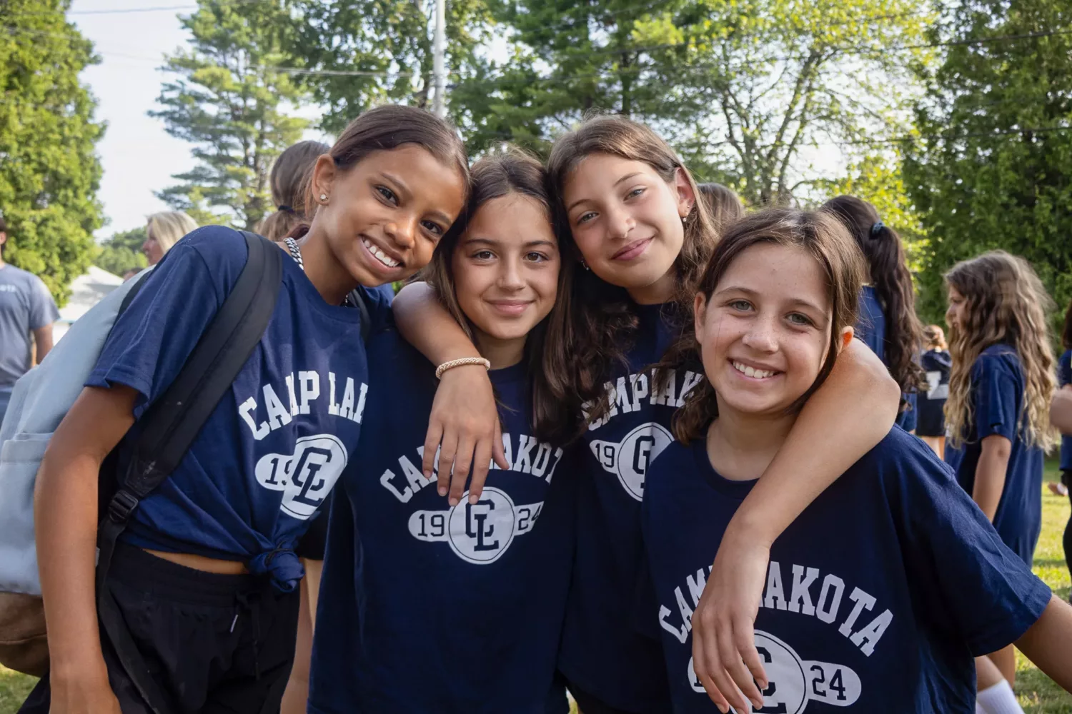 Four Camp Lakota campers in navy blue Camp Lakota t-shirts with arms around each other smiling outdoors