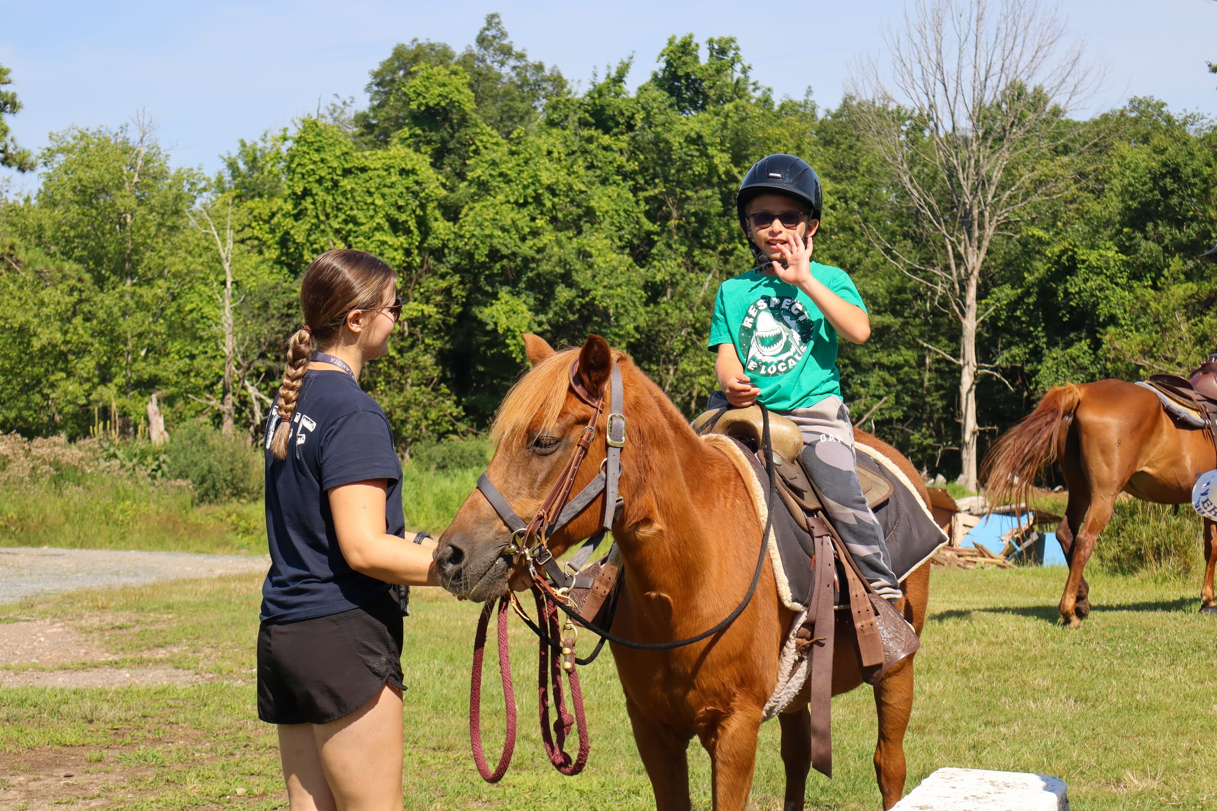 A child riding a horse and waving while an instructor holds the horse.