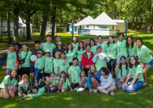 Large diverse group of Camp Lakota campers and counselors in green shirts posing on grass with camp buildings in background